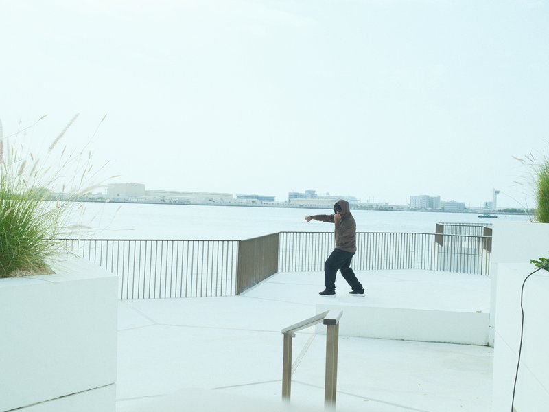 A person in a brown shirt gestures while standing on a white deck by a calm water body.