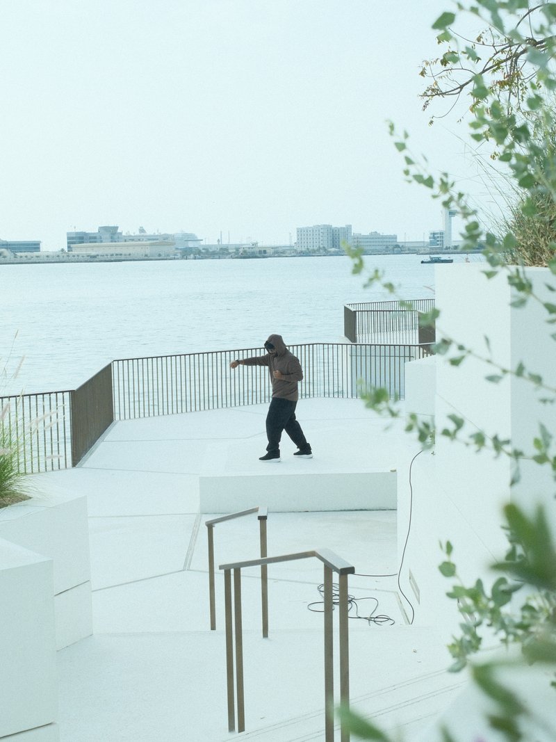 A person walking on a white terrace by the water, with a city skyline in the background.