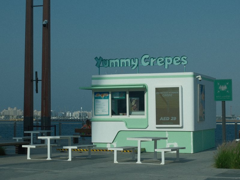 A small crepe stand with a green and white color scheme, surrounded by outdoor seating and a waterfront view.