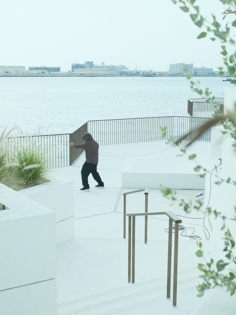 A person walking near a waterfront with modern architecture and greenery in the foreground.