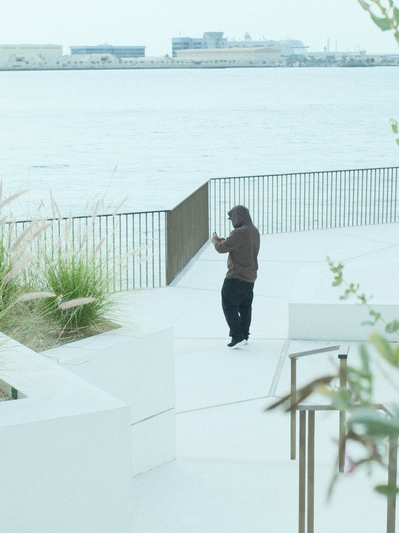 A person in a brown hoodie stands on a waterfront promenade, looking at their phone with a gray sky above.