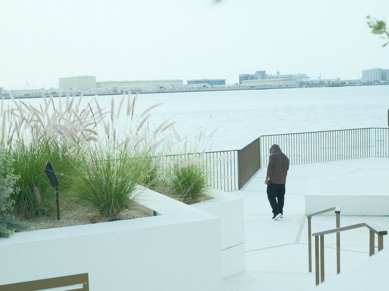 A person in a brown hoodie walks along a white pathway by a calm water body with distant buildings.