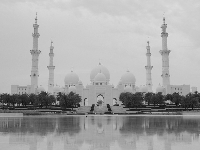 A grand mosque with multiple minarets reflected in a calm water body under a cloudy sky.