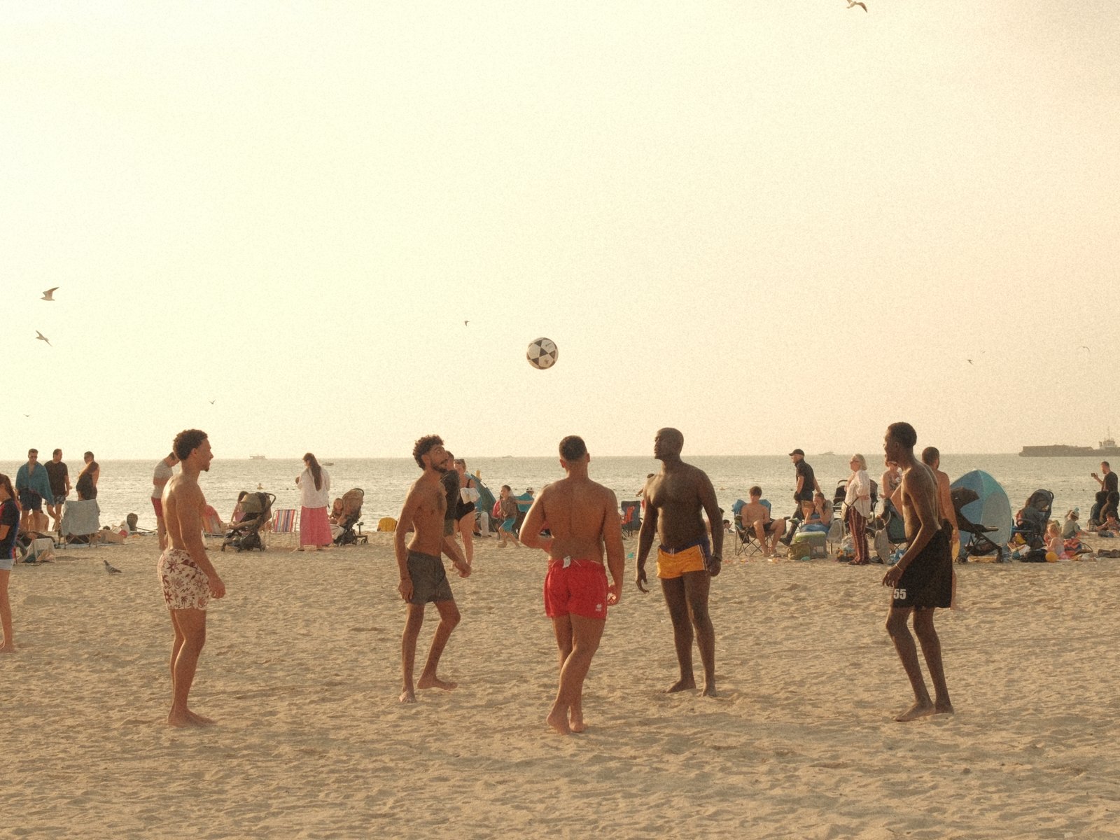 Group of people playing beach volleyball on a sandy beach with a hazy sky and distant beachgoers.