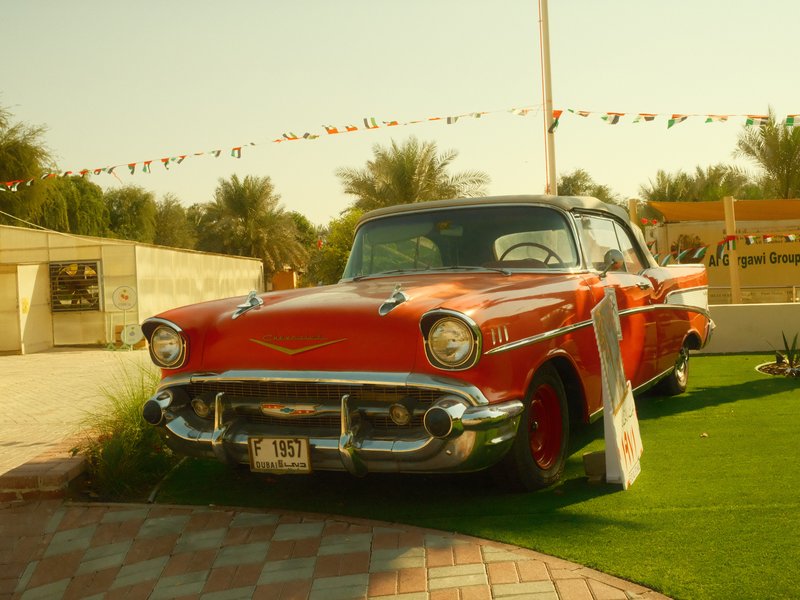 A vintage red car parked on green grass with palm trees in the background under a clear sky.