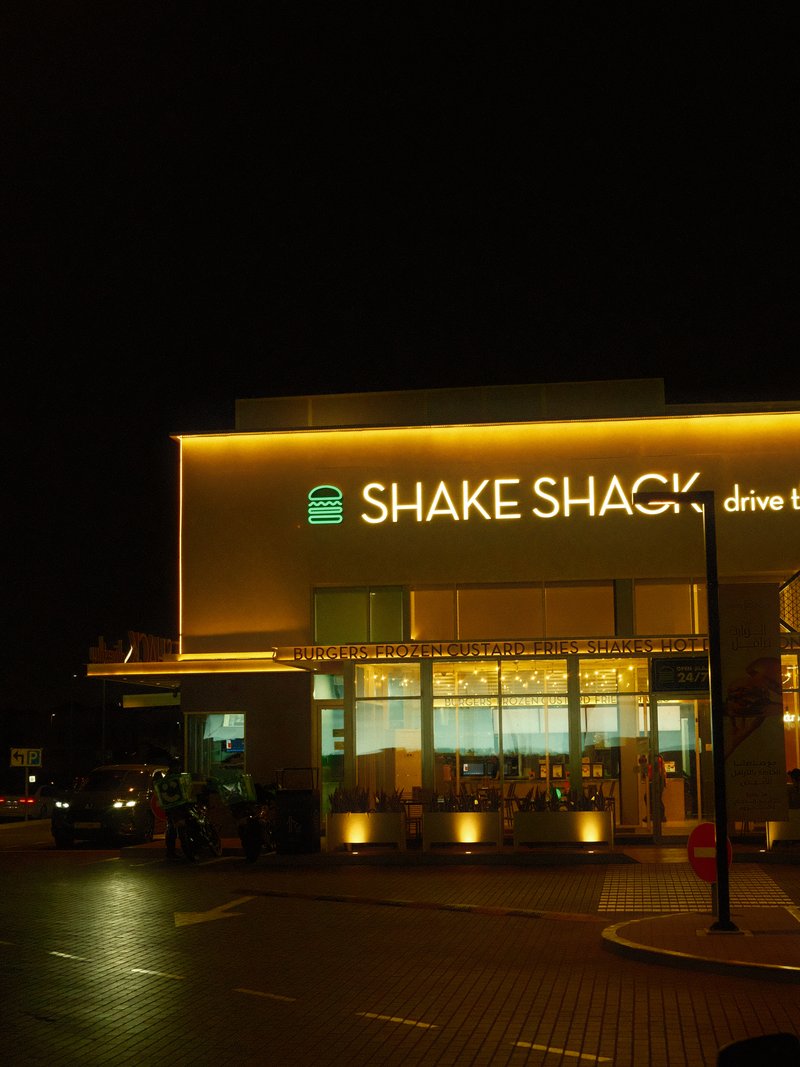 Exterior view of Shake Shack restaurant at night, illuminated sign and warm lights visible.