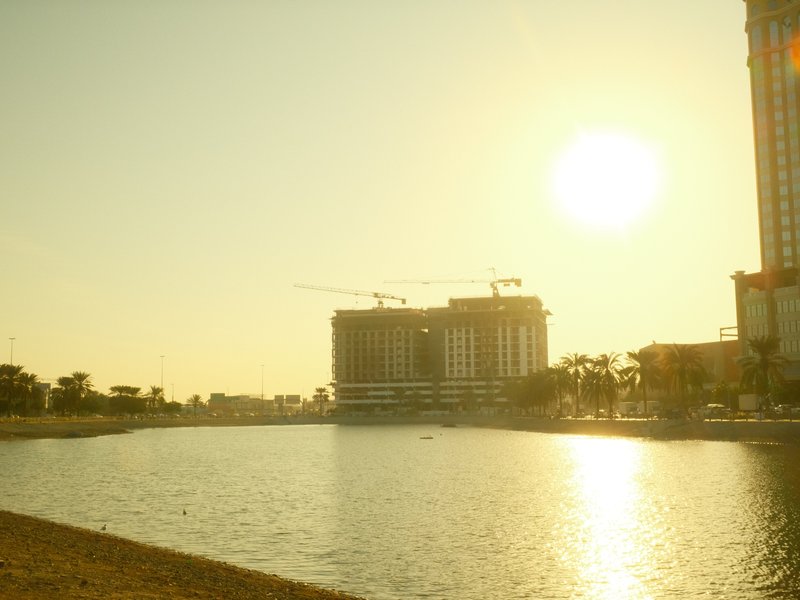 A serene lakeside view at sunset with buildings and cranes silhouetted against the golden sky.