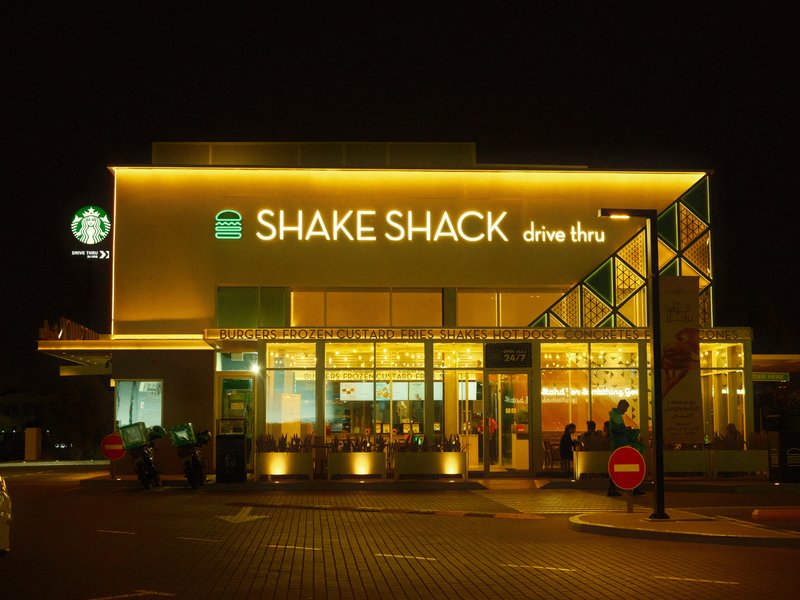 Exterior view of a Shake Shack restaurant illuminated at night with a Starbucks nearby.