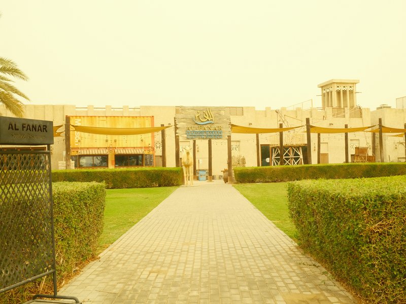 Entrance to a building with a pathway lined by hedges, under a yellowish light.