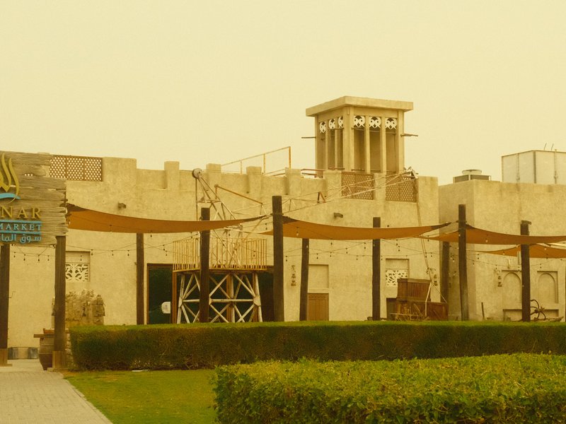 A historic building with a tower, surrounded by a green lawn and wooden structures under a yellowish sky.