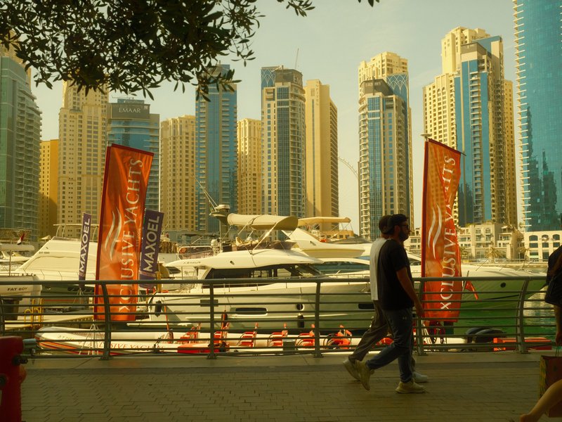 People walking along a waterfront with boats and tall buildings in the background under a warm light.