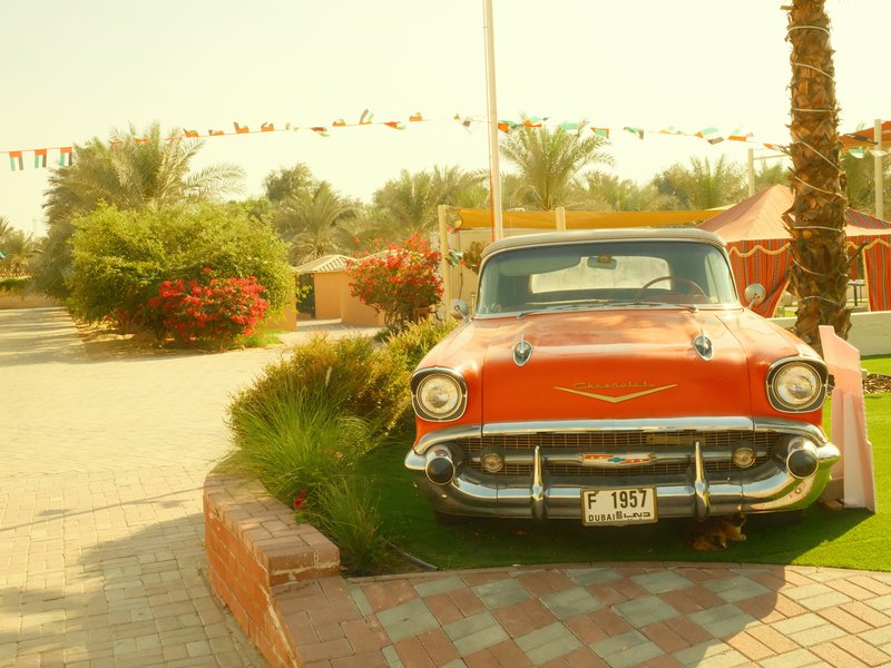 A vintage red car parked on a green lawn with palm trees and colorful decorations in the background.