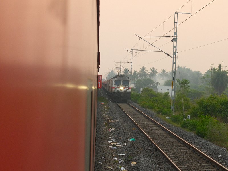 A train approaching on parallel tracks with a blurred red train in the foreground and greenery in the background.