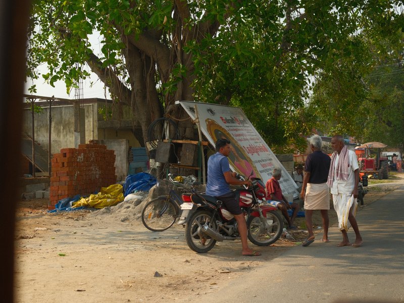 A street scene with people walking near a motorcycle and a makeshift stall under a tree.