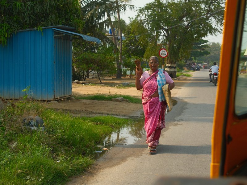A woman in a pink sari walking along a rural road, waving with a bag over her shoulder.