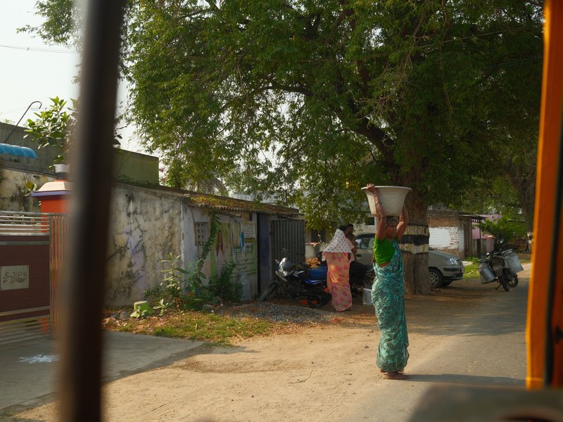 A woman carrying a pot on her head walks along a rural road lined with trees and buildings.