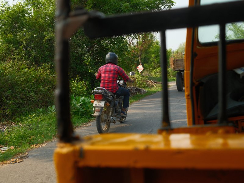 A motorcyclist in a red checkered shirt riding on a rural road, viewed through a vehicle's window.