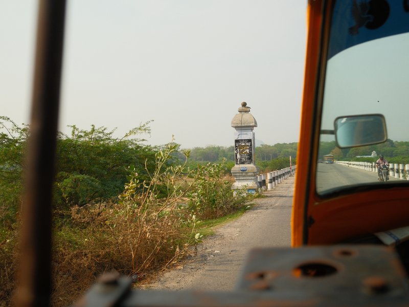 View from a vehicle showing a distant monument on a road surrounded by greenery.