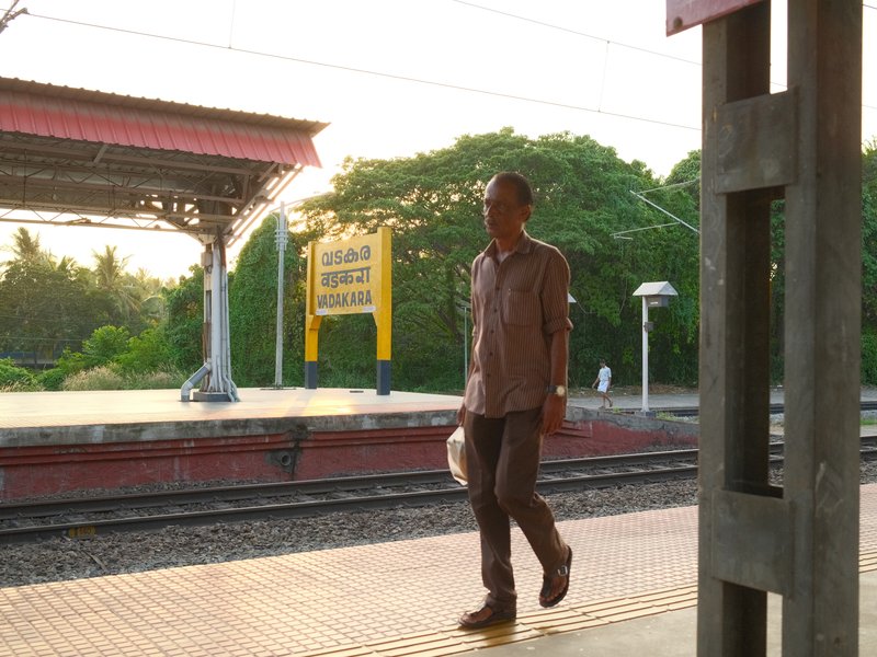 A man walking on a train platform during sunset, with trees and a station sign in the background.