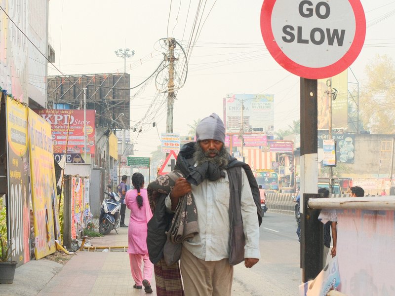 A man carrying a dog walks along a busy street with a 'GO SLOW' sign in the foreground.