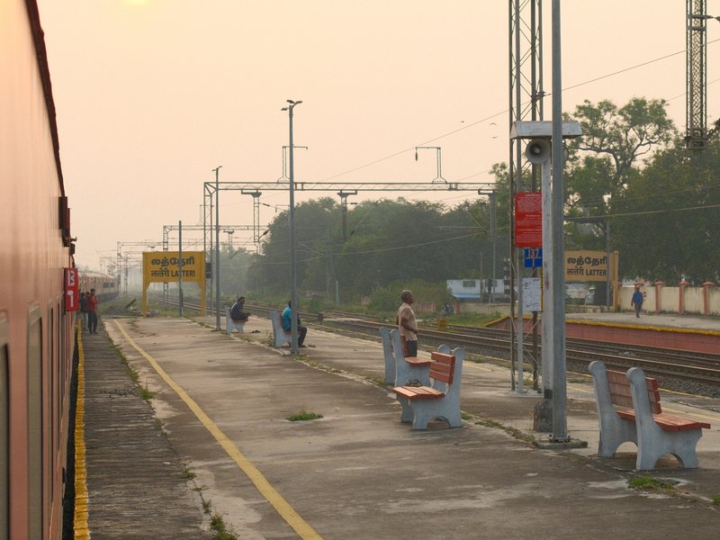 Train station platform with benches, people waiting, and a train in the background during a hazy evening.