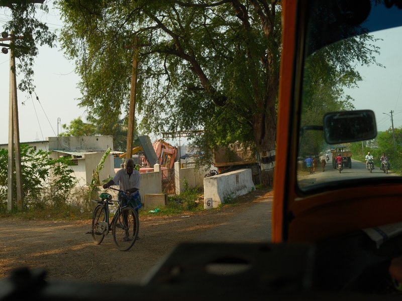 A boy with a bicycle stands on a rural road, surrounded by greenery and distant buildings.