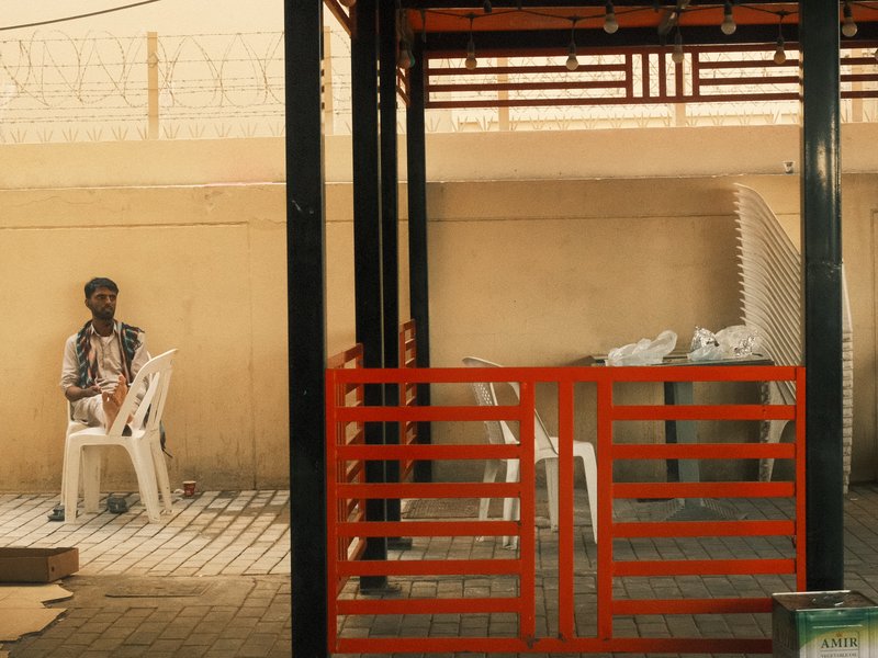 A man sitting on a plastic chair in a shaded area with orange railings and a beige wall.