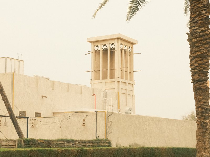 A traditional wind tower stands against a beige sky, surrounded by palm trees and a stone wall.