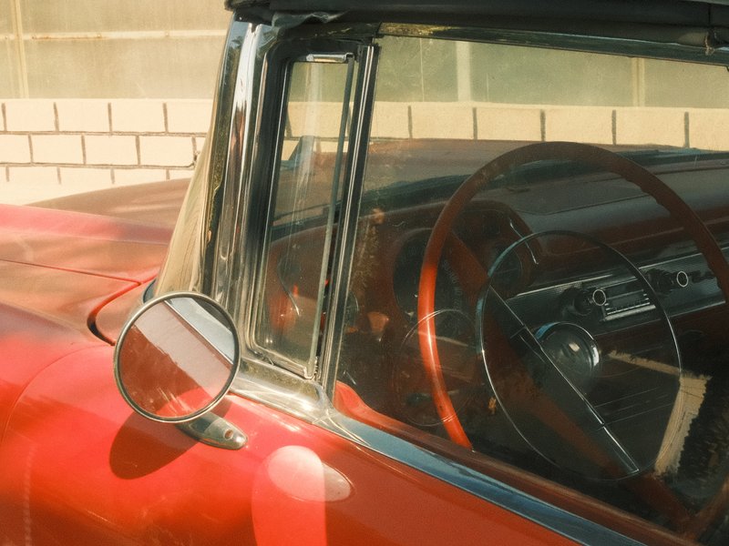 Close-up view of a vintage red car's interior showing the steering wheel and side mirror.