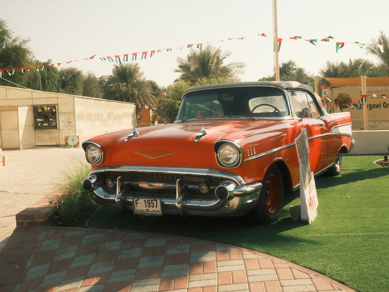 A vintage orange car parked on a green lawn with palm trees in the background.