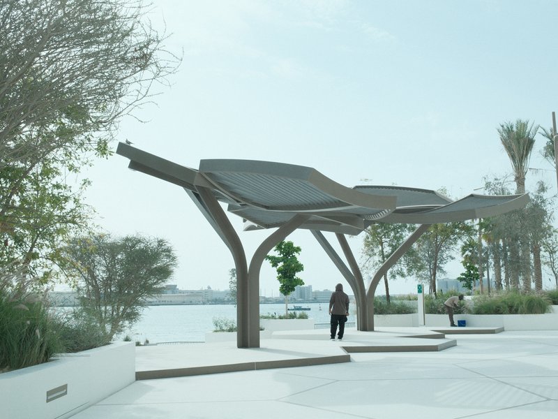 A modern pavilion structure by the water with a person walking underneath, surrounded by greenery.