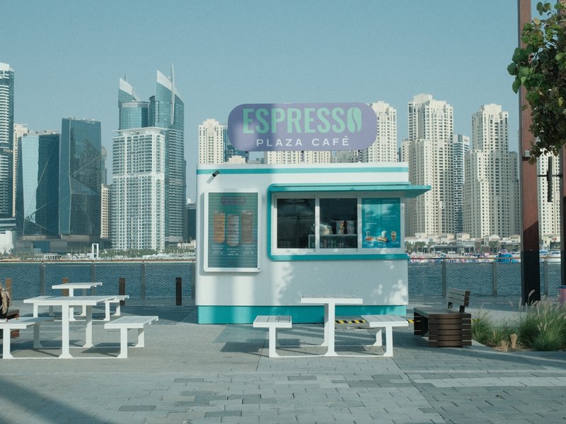 A small espresso kiosk by the waterfront with skyscrapers in the background under a clear sky.