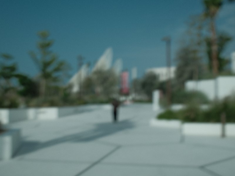A blurred view of a walkway with a person walking in an urban setting, surrounded by greenery and modern architecture.
