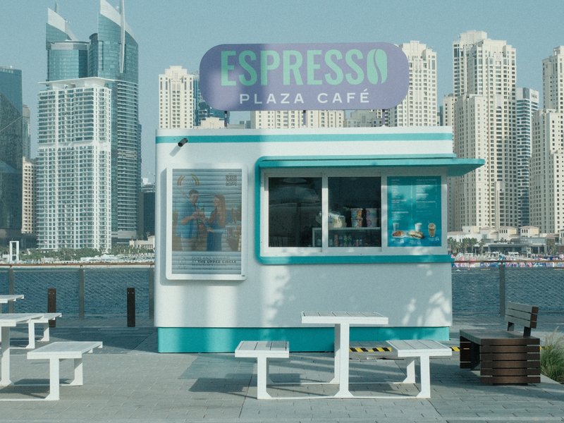 A small espresso café kiosk with a turquoise and white color scheme, set against a city skyline.