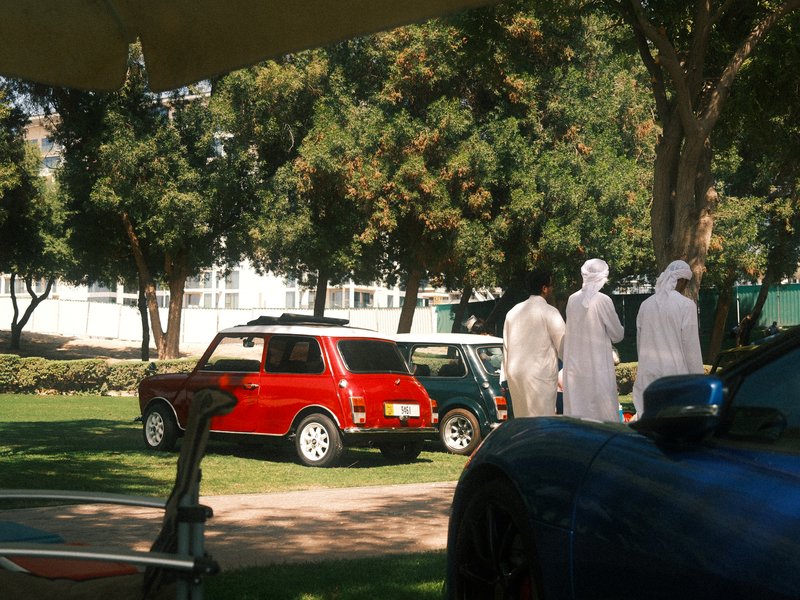 Two figures in white robes walking past classic cars in a park setting with trees.