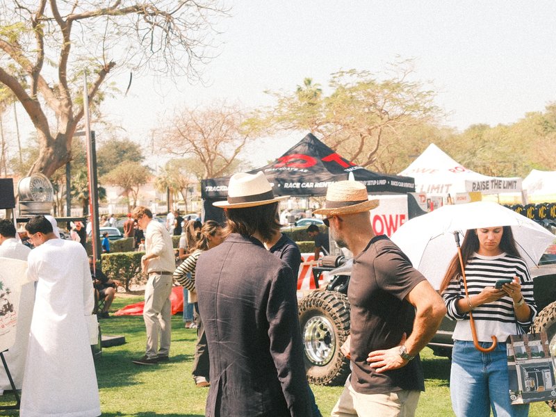 Two men in hats converse at an outdoor event with tents and people in the background.
