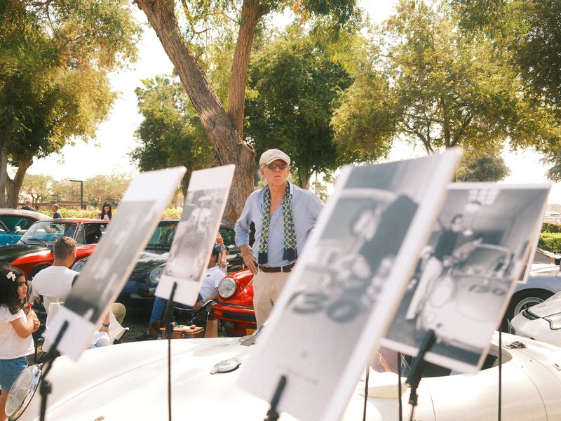 A man stands among displayed photographs at an outdoor car show, surrounded by trees and vintage cars.