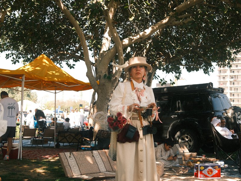 A woman in a white outfit stands under a large tree at an outdoor event, with tents and people in the background.