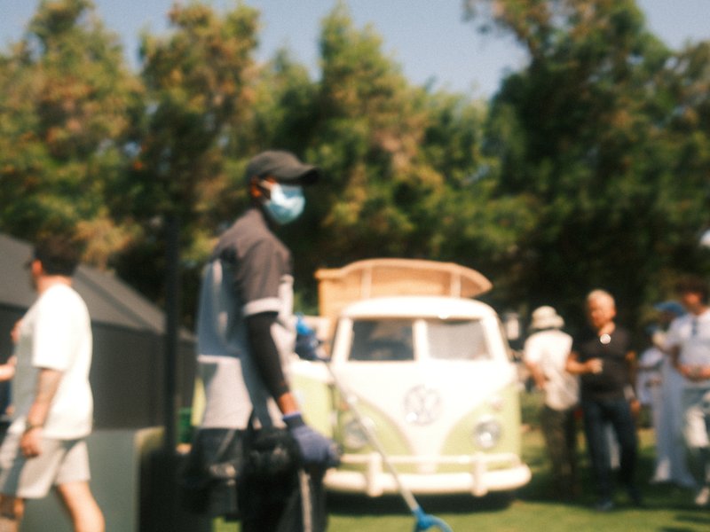 A man in a mask stands with a shovel near a vintage van, surrounded by people in a sunny outdoor setting.