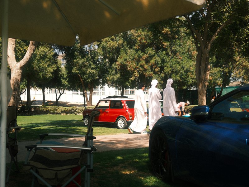 A red car parked on a grassy area with people in light clothing walking in the background under trees.