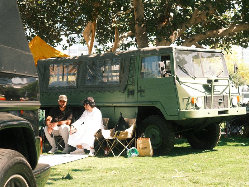 Two people sitting on chairs next to a military-style green vehicle in a grassy area.