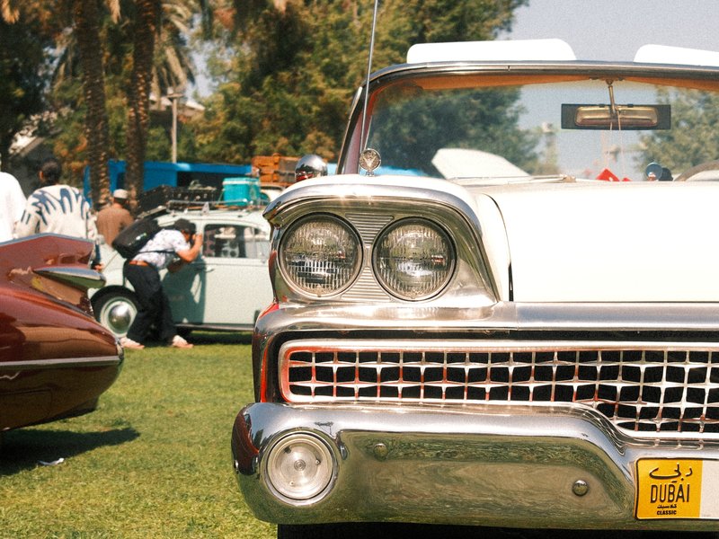 Close-up of a classic white car in a grassy area with vintage cars and people in the background.