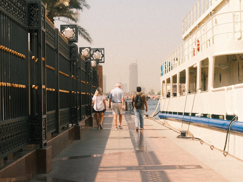Three people walking along a waterfront promenade beside a boat, with palm trees and a city skyline in the background.
