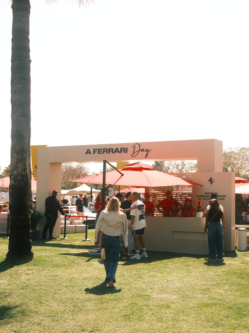 People enjoying a Ferrari Day event with red umbrellas and a white booth in a sunny outdoor setting.