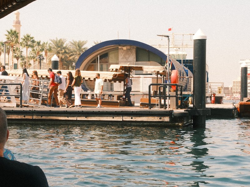 People waiting at a dock near water with palm trees and a building in the background.