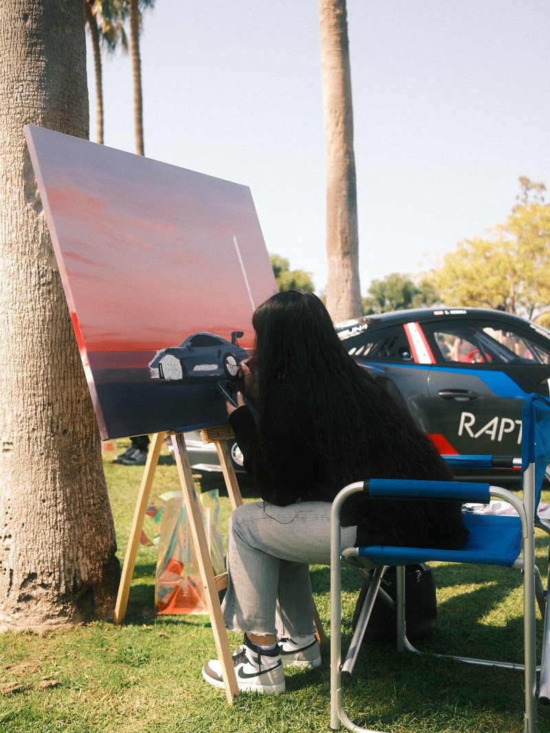 A person painting a car on a canvas in a park, with palm trees and a race car in the background.