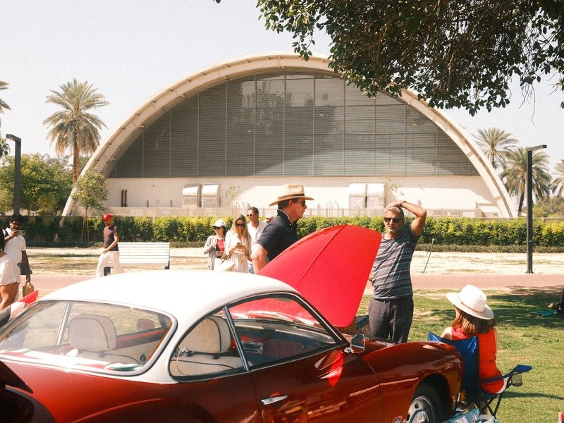 A vintage red car in the foreground with people interacting and a large modern building in the background.