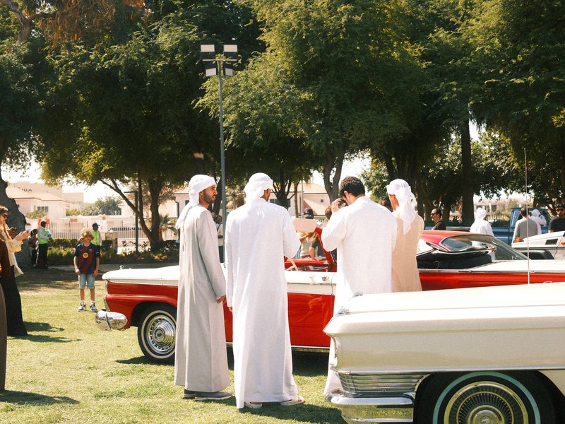 Group of men in traditional attire gathered around a classic red convertible car in a park.