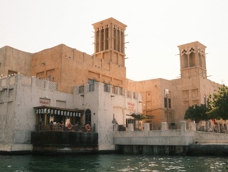 Historic building with wind towers by the water, people enjoying the view under warm sunlight.