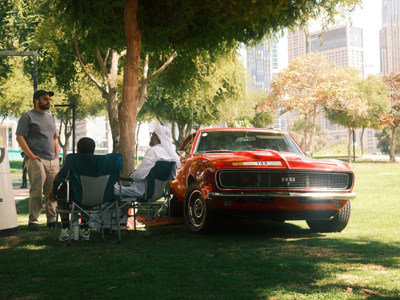 A classic red car parked in a park with people sitting nearby, surrounded by trees and city buildings.